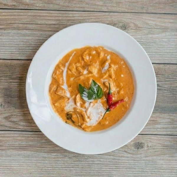 Orange curry with chicken, basil leaves, and red chili peppers in a white bowl.