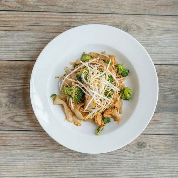 A white plate of Pad See Ew with flat noodles, broccoli, chicken, and bean sprouts on a wooden table.