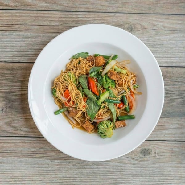 Stir-fried noodles with tofu, broccoli, green beans, red peppers, and basil in a white bowl.
