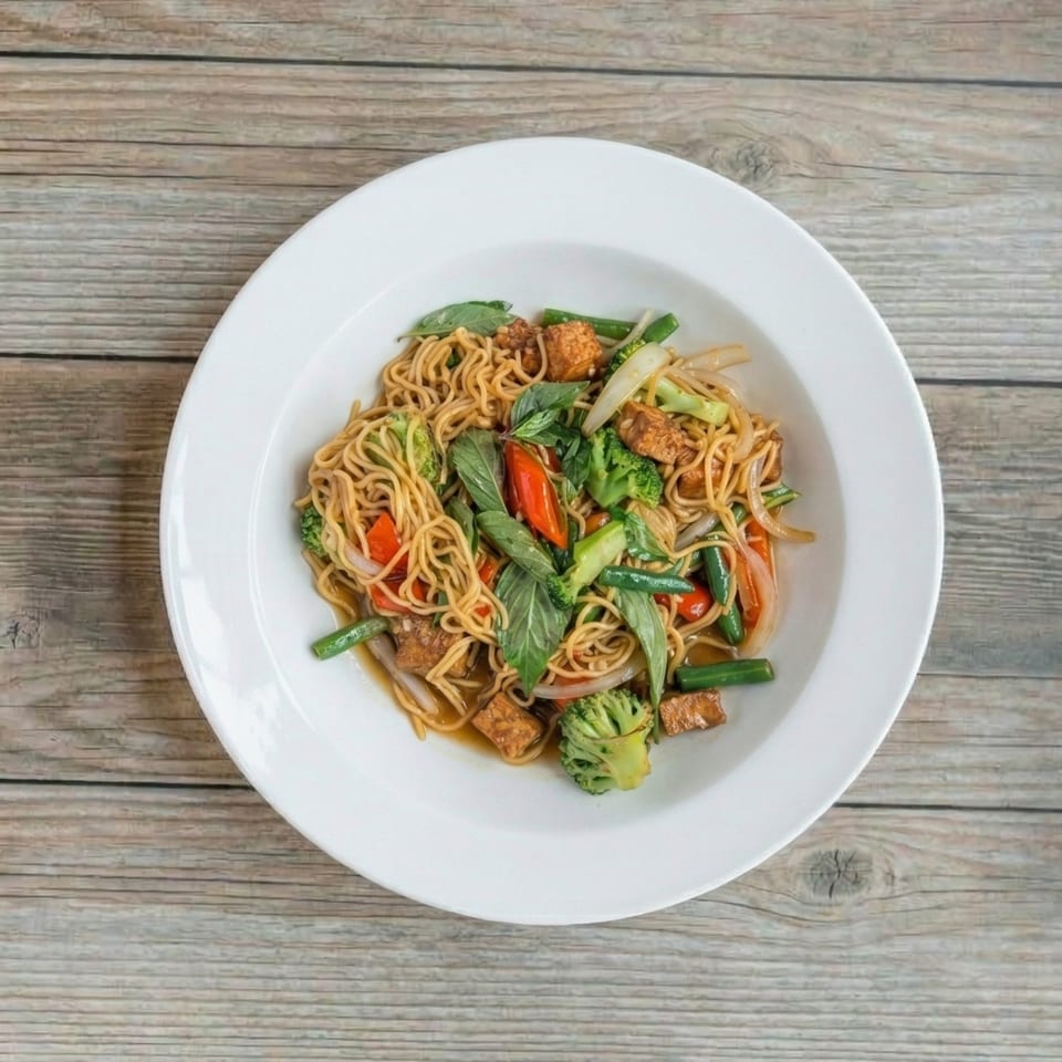 Stir-fried noodles with tofu, broccoli, green beans, red peppers, and basil in a white bowl.