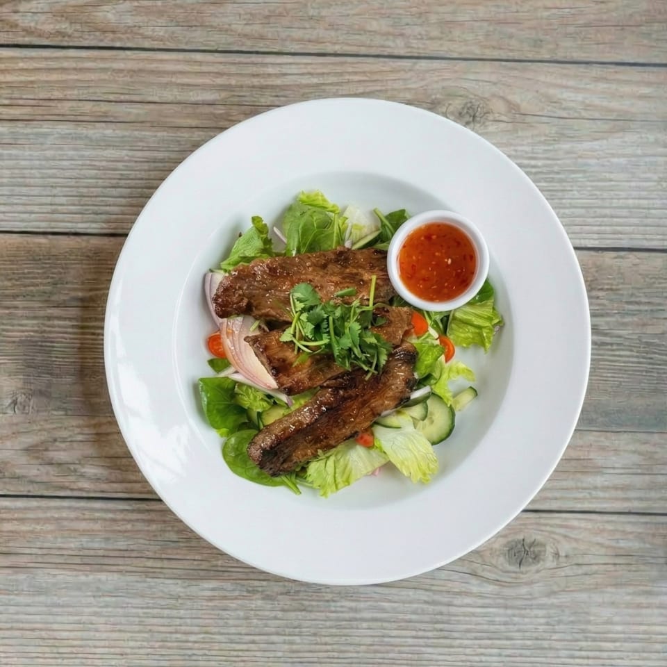 Grilled beef salad with mixed greens, cucumbers, red onions, tomatoes, cilantro, and a side of chili sauce.